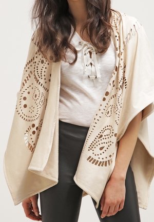 Frau trägt einen beigefarbenen Poncho mit aufwendigen Ausschnittmustern über einem weißen Spitzen-Top mit Schnürung und schwarzen Hosen.