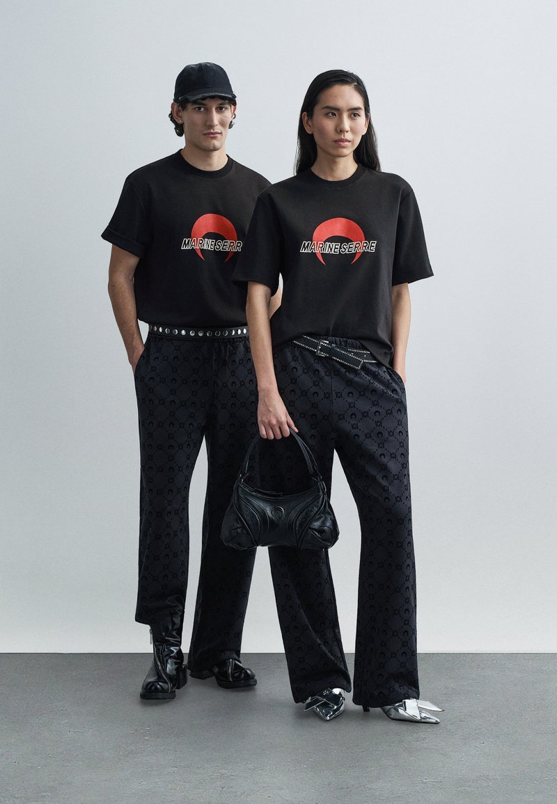 Δύο μοντέλα στέκονται δίπλα-δίπλα φορώντας μαύρα t-shirts Marine Serre και μαύρο παντελόνι με σχέδια· το ένα κρατά μια μαύρη τσάντα χειρός.