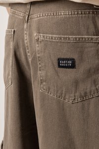 Braune Baumwollhose mit einer Gesäßtasche, die ein schwarzes rechteckiges Etikett mit der Aufschrift "KAOTIKO SOCIETY" trägt. Sichtbare Nähte entlang der Säume.