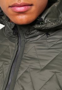 Detailní záběr na osobu v olivově zelené prošívané bundě se zipem vpředu a nastavitelnými šňůrkami kapuce u brady.