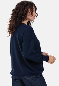 Pull en maille bleu marine à coupe décontractée, avec un col rond et des poignets côtelés, associé à un jean en denim foncé. Tissu texturé visible.