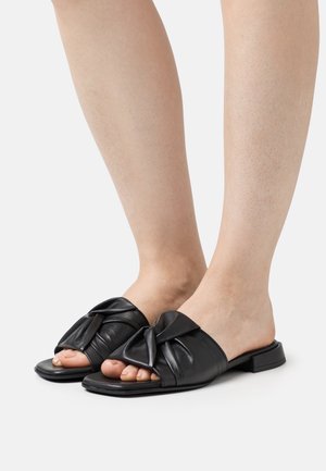 Schwarze Leder-Sandalen mit einem geknoteten Design an der Zehe, flacher Sohle und offenem Rücken, der die Zehen zeigt. Glatte Textur.
