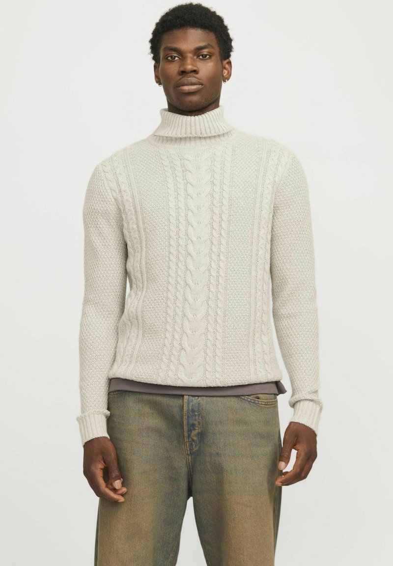 Suéter de cuello alto gris claro con patrones de punto de cable texturizados y puños acanalados, combinado con jeans holgados de color azul.