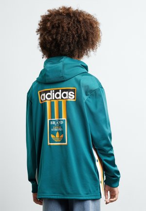 Person med krøllet hår vendt væk, iført en teal Adidas hættetrøje med gule striber og logo på ryggen mod en ensfarvet baggrund.