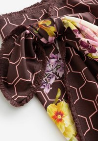 Brūns šalle ar rozā sešstūra rakstu, bārkstīm malās un ziedu motīviem dzeltenā, violetā un zaļā krāsā, vaļīgi sasieta.