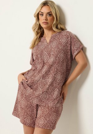 Kvinde med bølget blondt hår iført et løst sæt bestående af en vinrød top og shorts med hvide geometriske mønstre, stående op ad en hvid væg.