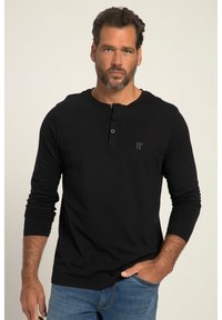 Schwarzes Langarmshirt aus Baumwolle mit einem Henley-Ausschnitt und drei Knöpfen, sowie einem kleinen Logo auf der Brust. Kombiniert mit blauen Jeans.