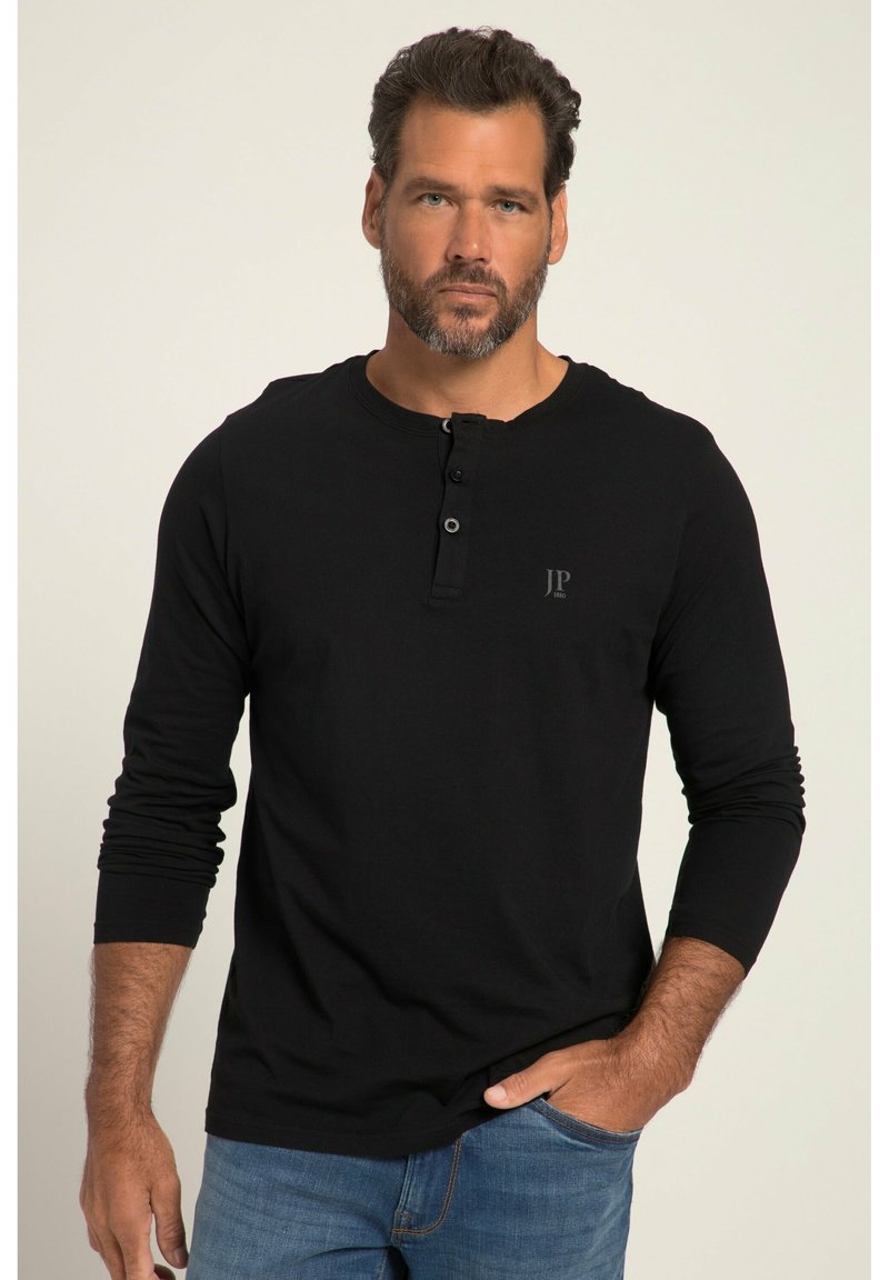 Schwarzes Langarmshirt aus Baumwolle mit einem Henley-Ausschnitt und drei Knöpfen, sowie einem kleinen Logo auf der Brust. Kombiniert mit blauen Jeans.