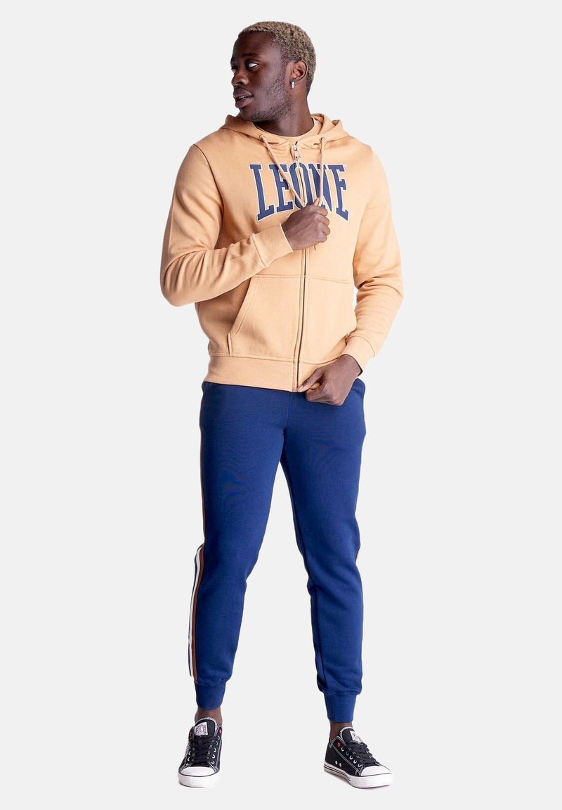 Homme portant un sweat à capuche beige avec fermeture éclair et le texte "LEONE", un pantalon de jogging bleu marine avec des bandes latérales et des baskets basses noires, debout et regardant à gauche.