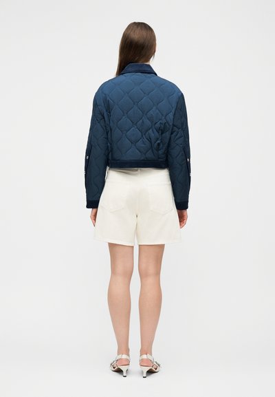 Femme portant une veste matelassée bleu marine, un short blanc et des sandales blanches à talons, debout de dos sur un fond clair uni.