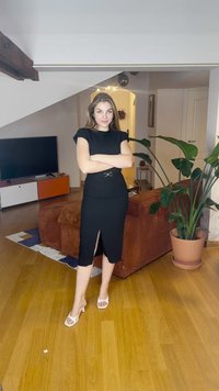 Crna uska haljina s kratkim rukavima, bočni prorez i ukrasni remen. Uparena s bijelim sandalama na petu na drvenom podu.