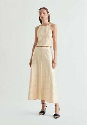 Mujer delgada con cabello largo y castaño que lleva un top corto con cuello halter beige texturizado y falda de talle alto a juego con sandalias negras de tacón.