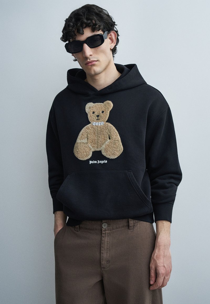 Jonge man met krullend haar, zwarte zonnebril, zwarte hoodie met teddybeerprint en bruine broek, staand tegen een effen grijze achtergrond.