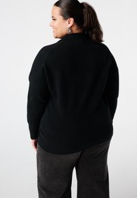 Zwarte gebreide trui met een ontspannen pasvorm en geribbelde details, gedragen met grijze wijde pijpen broek. Achteraanzicht toont een gladde, gestructureerde stof.