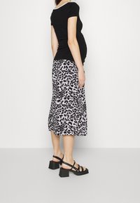 Donna incinta in top nero aderente, gonna midi con stampa leopardata e sandali neri con tacco, in piedi contro uno sfondo chiaro e semplice.