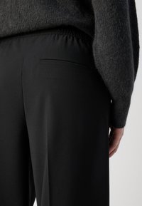 Black trousers with a smooth texture, elastic waistband, and a single back pocket. Paired with a dark, fuzzy sweater.