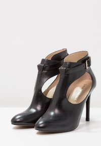 Černé kožené kotníkové boty na vysokém podpatku se vykrojenými stranami, hladkým povrchem, špičatou špičkou a zlatou sponou jako akcentem.