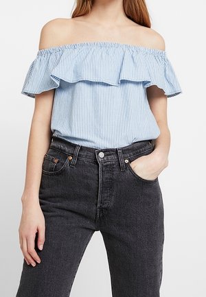 Vrouw draagt lichtblauw gestreepte off-shoulder blouse, ingestopt in high-waisted zwarte denim jeans, één hand in de zak.