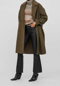 Nadrozměrný vlněný kabát olivově zelené barvy, vzorovaný pletený svetr v přírodních tónech, černé kožené kalhoty do zvonu a špičaté černé kotníkové boty.