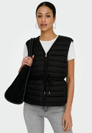 Vrouw draagt een zwarte gewatteerde mouwloze jas over een wit shirt met korte mouwen, lichtgrijze spijkerbroek, en houdt een grote zwarte schoudertas vast.