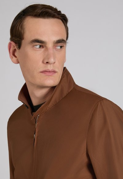 Giovane uomo con capelli castani corti che indossa una giacca marrone chiusa con la zip, guarda verso sinistra su uno sfondo chiaro e uniforme.