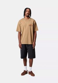 Camiseta de gran tamaño color beige con un logo en el pecho, combinada con shorts de mezclilla negros a la altura de la rodilla, calcetines blancos y zapatos marrones con cordones.