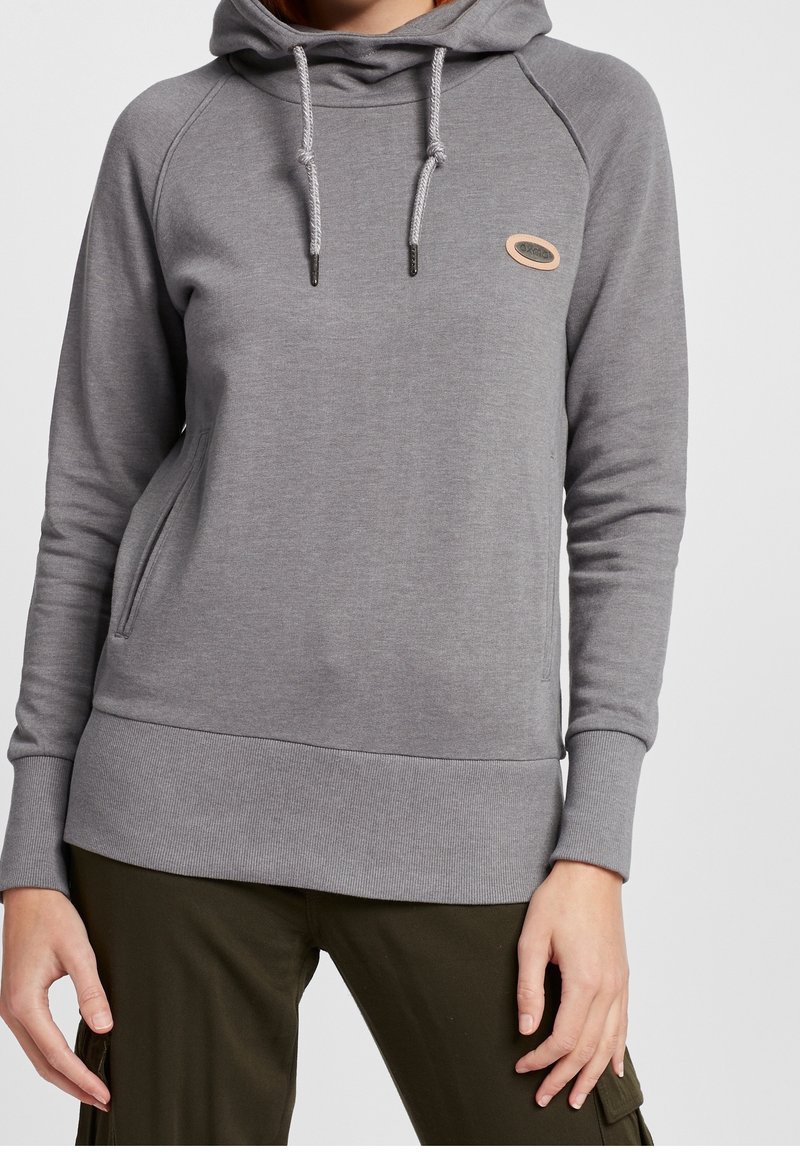 Personne portant un sweat à capuche gris avec des poches avant et un pantalon vert foncé, debout devant un fond clair uni.