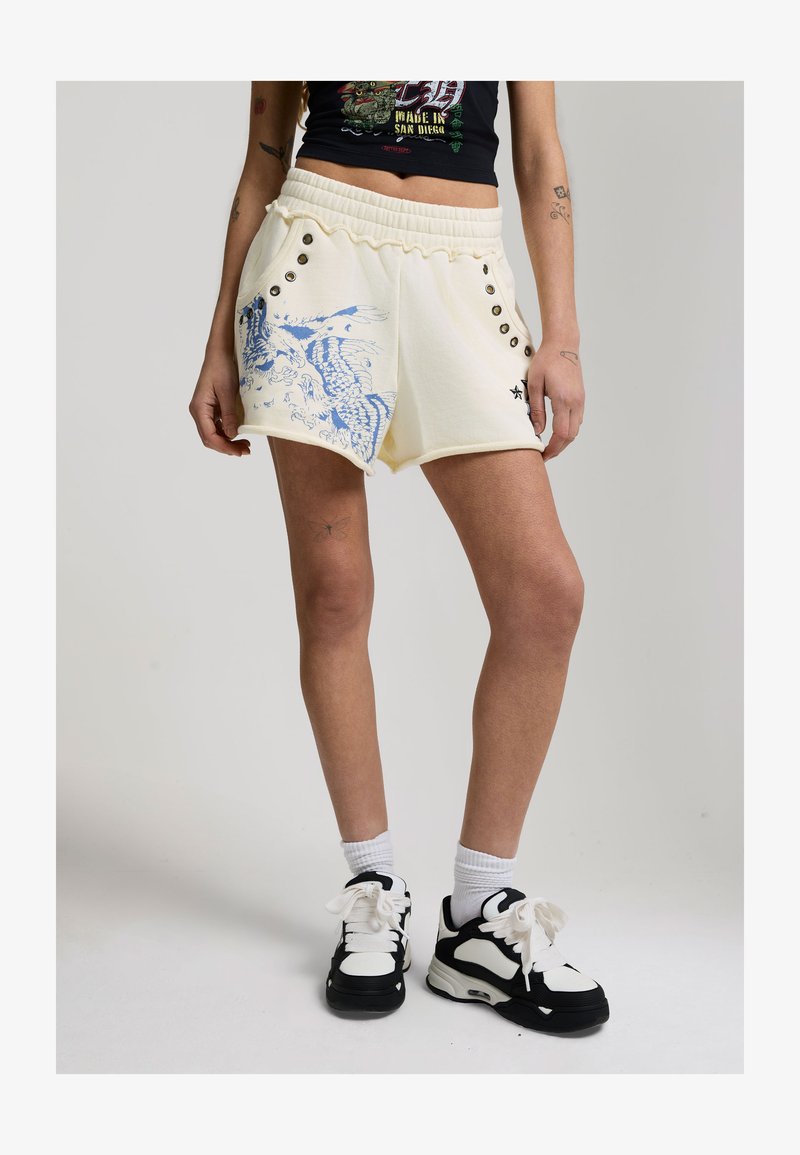 Vrouw die crèmekleurige short draagt met blauwe draakprint en metalen oogjes, zwart-witte sneakers, en een zwart cropped top met grafische print.
