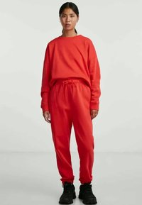 Rød, teksturert hettegenser med bred halsing og matchende joggebukser med elastisk linning og snor. Svarte, kraftige joggesko.