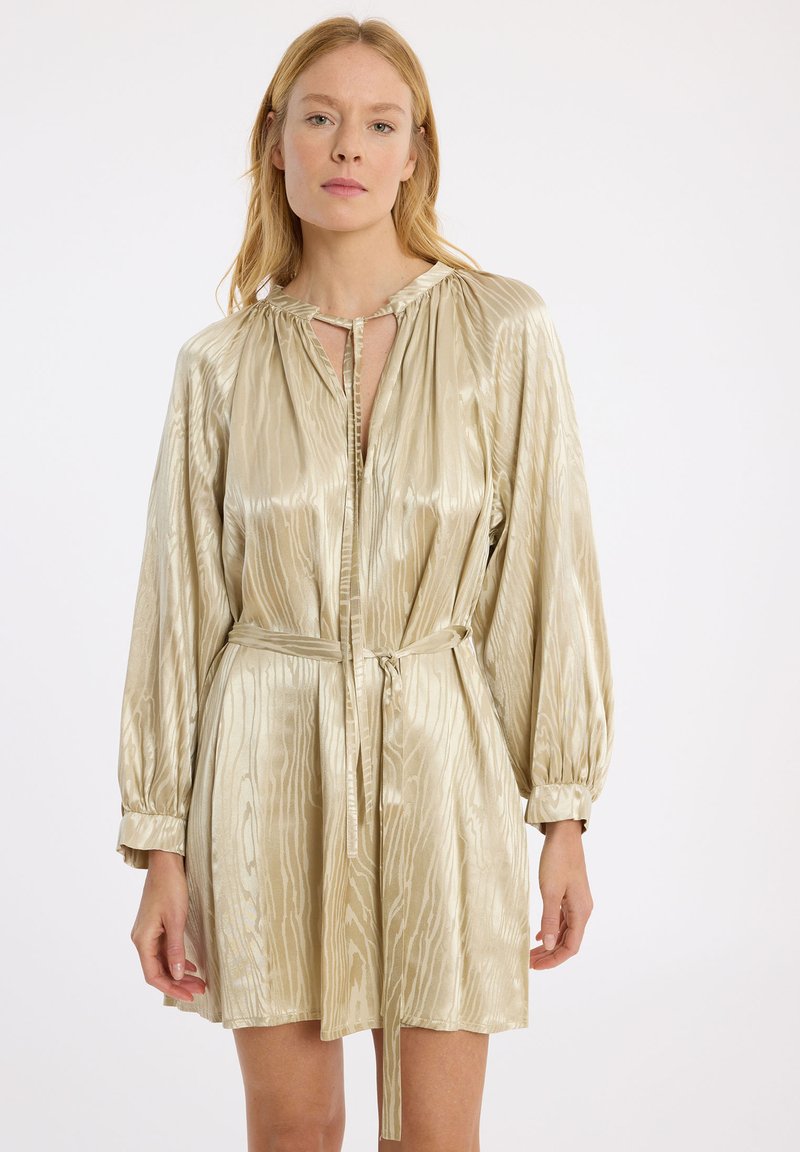 Robe dorée courte avec une taille à nouer, de longues manches ballon et un éclat texturé. Présente une fente au niveau du décolleté et un motif rayé subtil.