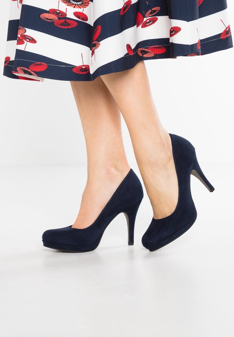 Jambes portant des escarpins en daim bleu marine associés à une jupe blanche avec des rayures bleu marine et des motifs floraux rouges.