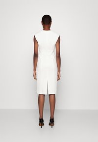 Vit ärmlös klänning med en figurnära siluett, ryggslits och slät textur, i kombination med svarta högklackade skor.