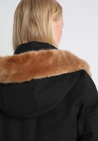 Czarna kurtka z odpinanym brązowym futrem sztucznym, posiadająca kaptur i widoczne detale szwów na materiale.
