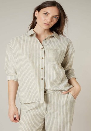 Femme portant une chemise boutonnée à rayures verticales beige et blanches avec poche poitrine et un pantalon assorti, debout avec une main dans la poche.