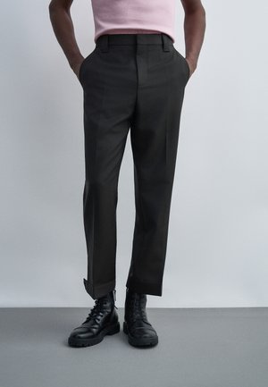 Personne portant un pantalon noir sur mesure avec revers et des bottes noires à lacets, mains dans les poches, sur un fond gris clair uni.