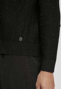Pull en tricot noir avec un motif texturé, comportant un ourlet et des poignets côtelés, et un accent de logo en métal rond en bas.