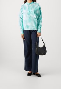 Un hoodie tie-dye turchese con tasca a marsupio, abbinato a pantaloni larghi blu navy e una borsa nera a forma curvata. Le scarpe in pelle slip-on completano l'outfit.