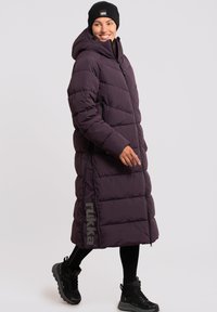 Lang lilla puffjakke med hætte, med lynlås lukning, sidelommer og et gråt "rukka" logo på siden. Bæres med sorte leggings og støvler.