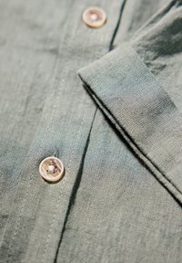 Camisa de lino verde con cuello de punta, tejido texturizado y un botón redondo de color claro en el frente.