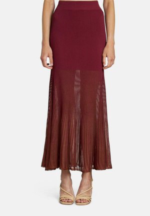 Falda maxi plisada burdeos con una cinturilla acanalada. La parte inferior transparente crea una silueta fluida. Un par de sandalias nude con tiras son visibles.