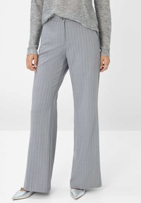 Grijze pinstriped broek met een getailleerde pasvorm, platte voorkant en wijde pijpen. De stof heeft een gladde structuur. Gedragen met een lichtgrijze trui en zilveren schoenen.