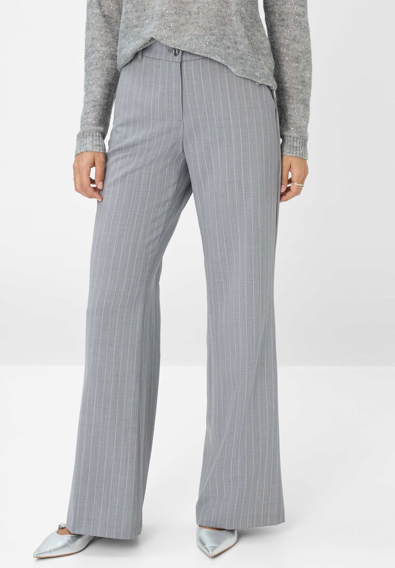 Grijze pinstriped broek met een getailleerde pasvorm, platte voorkant en wijde pijpen. De stof heeft een gladde structuur. Gedragen met een lichtgrijze trui en zilveren schoenen.