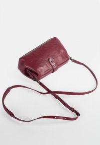 Burgundy leather crossbody bag with a smooth texture, rounded shape, adjustable strap, and snap closure detail. Minimalist design.