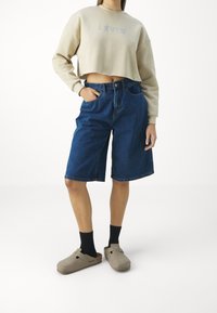 Crop sweatshirt crema con logo "LEVI'S", abbinato a pantaloni Bermuda in denim blu. Ciabatte grigie indossate con calze nere. Design semplice.