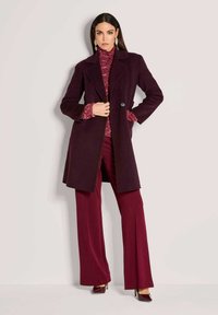 Vrouw in een donkerpaars jack over een bordeauxrode trui en wijde bordeauxrode broek, poseert met haar hand in de zak van het jack tegen een effen muur.