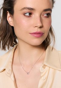 Srebrny naszyjnik z małym, okrągłym, przezroczystym zawieszeniem z kamieniem szlachetnym. Noszony na jasnobeżowej satynowej koszuli z delikatnym połyskiem.