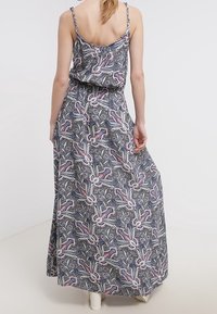 Kobieta z blond włosami ubrana w bezrękawnikową, wzorzystą sukienkę maxi w różowo-niebieskie kwiatowe wzory, stojąca na tle jasnego, jednolitego koloru.