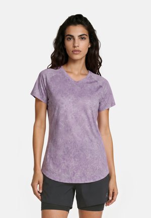 Vrouw met lang donker haar die een paarse tie-dye korte mouwen V-hals T-shirt en zwarte sportshorts draagt, staand tegen een effen achtergrond.