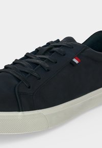 Baskets basses noires avec lacets noirs, semelle en caoutchouc blanche et une petite étiquette en tissu rouge, blanche et noire sur le côté.