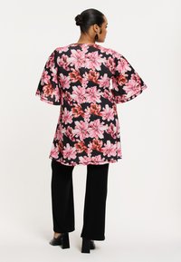 Bloemrijke tuniek met roze en rode bloemen op een zwarte achtergrond, met losse mouwen, gecombineerd met zwarte broeken en chunky hakken.